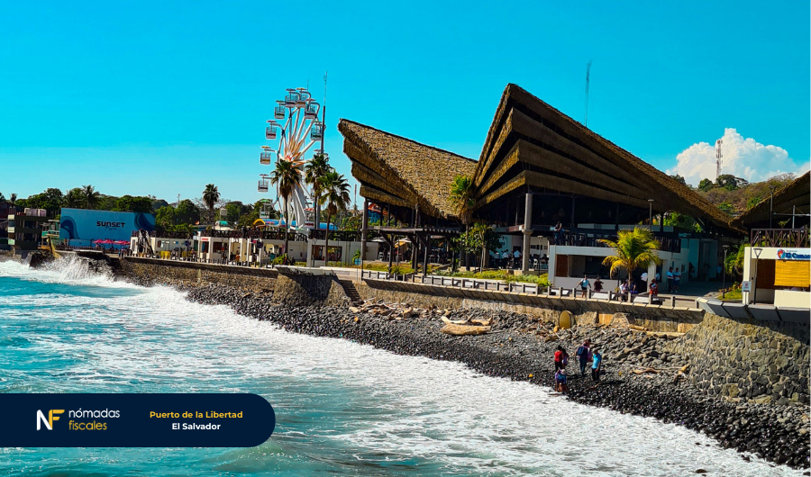 Vista del malecón de Puerto de La Libertad en El Salvador, con el mar rompiendo contra las piedras, una noria y un moderno centro turístico al fondo.